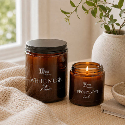 Two amber glass candles on a wooden table in a softly styled room. One candle is unlit with a black lid and reads 'WHITE MUSK and Amber'; the other is lit with a black rim and reads 'PEONY and SOFT Suede'.