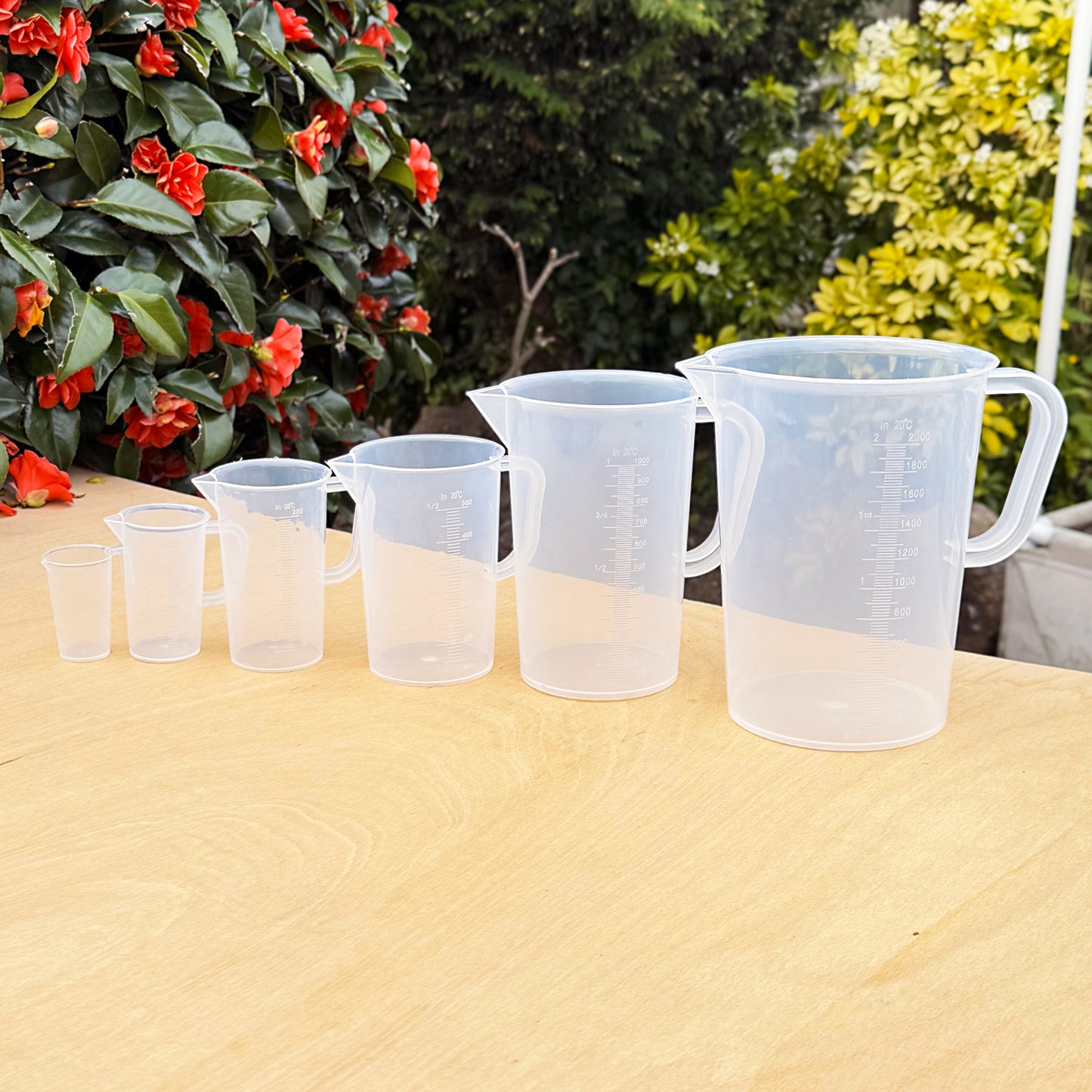Plastic measuring jug set – sizes from 50ml to 5L lined up on wooden surface with garden background, ideal for gardening, baking, and DIY